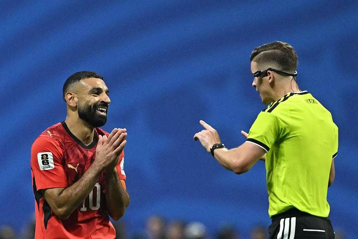 During a soccer match, a player in a red uniform is seen gesturing while another individual, dressed in a green shirt and wearing a whistle around their neck, engages in conversation with them. The background features a blue field and spectators in the stands.